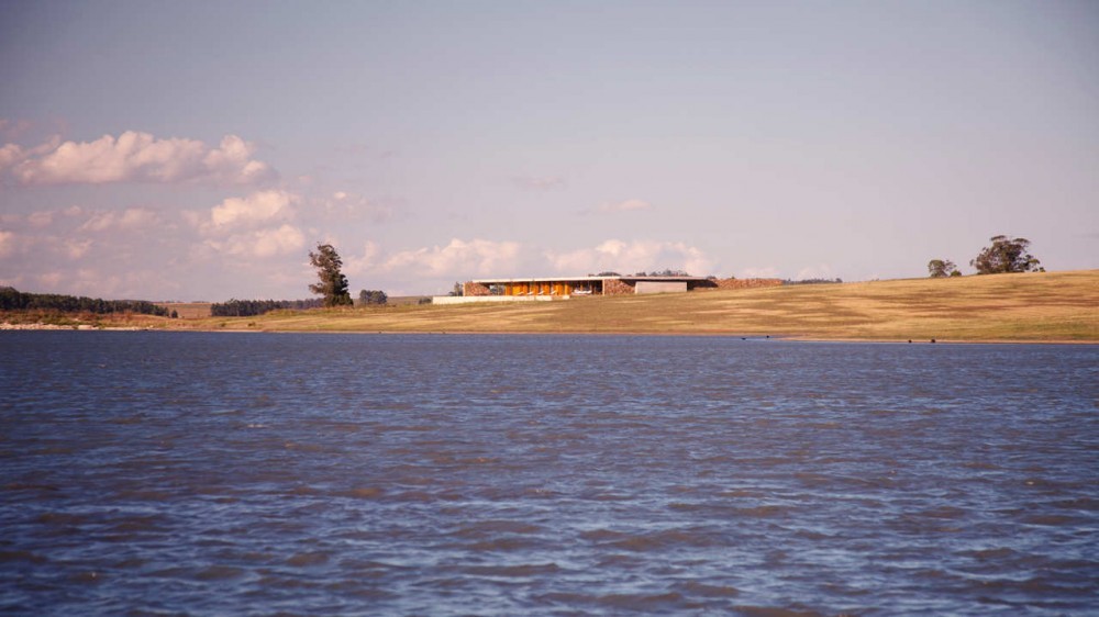 Low house with a flat roof on a grassy shoreline, seen across rippled lake water under scattered clouds