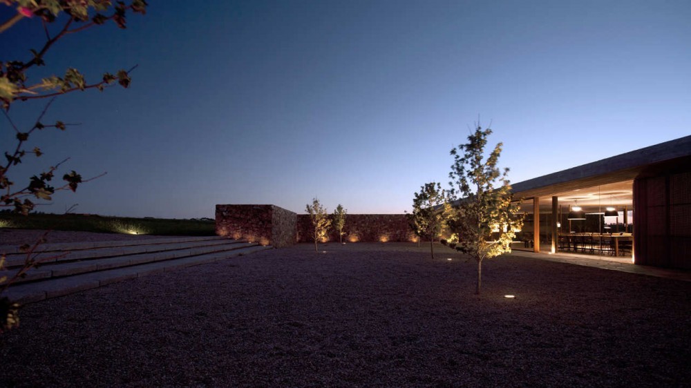 Gravel courtyard with wide stone steps, a rubble wall and lit saplings beside a glazed dining pavilion at dusk
