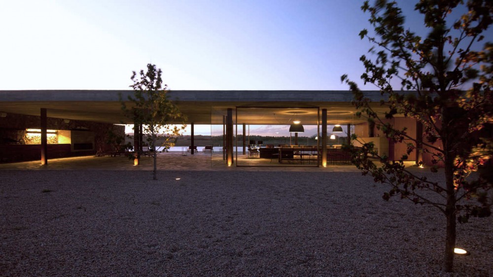 Glass-fronted living pavilion under a flat concrete roof, glowing across a gravel court with small ground lights at twilight