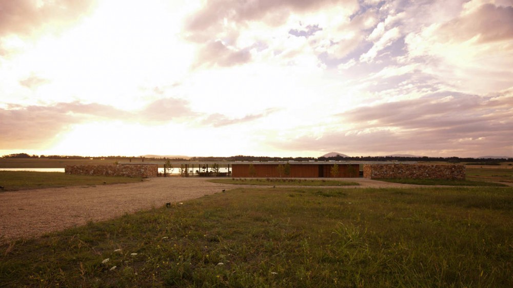 Distant low pavilion between two rough stone walls in open grassland under a glowing, cloudy sunset sky