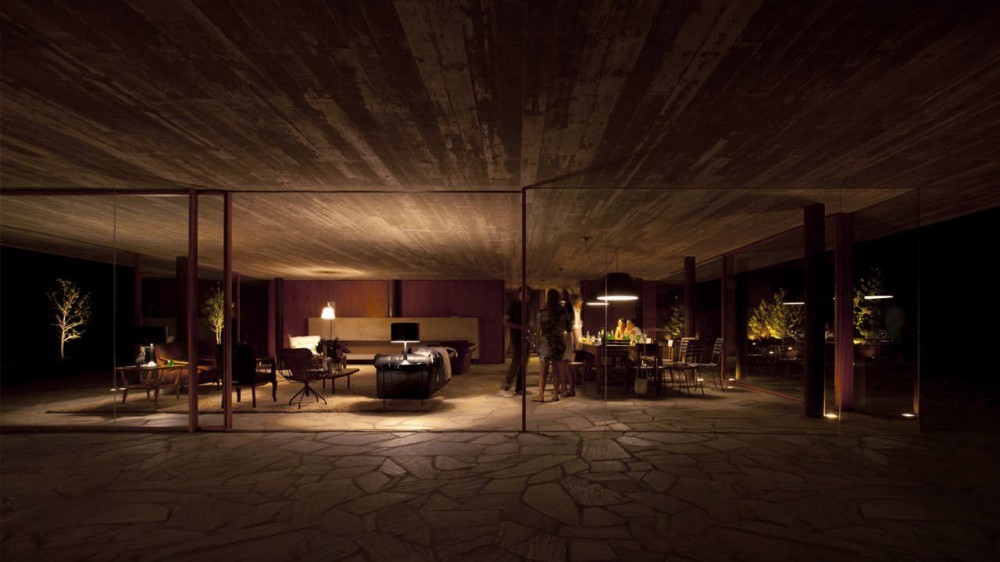 Night view through glass walls into a lit living and dining area, with people gathered beneath a concrete ceiling