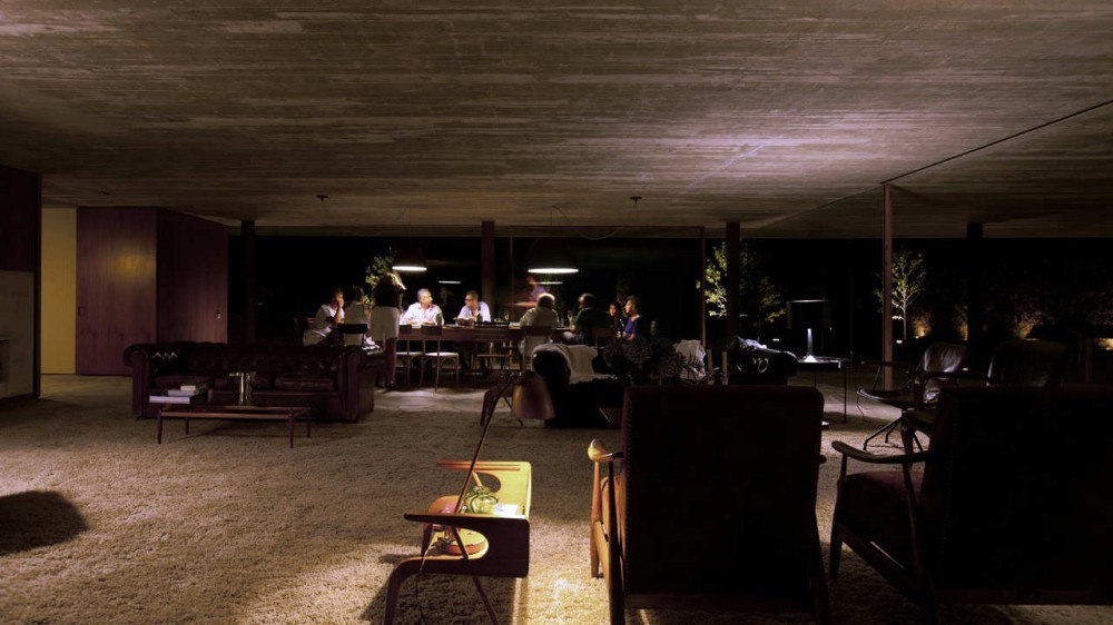 Dinner party seated at a long table beyond sofas and armchairs, under a board-marked concrete ceiling at night