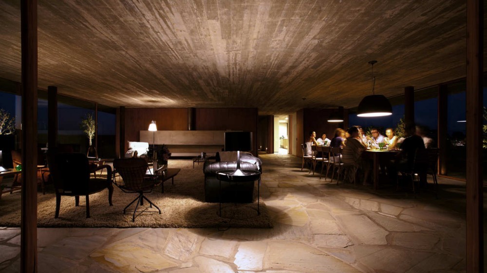 Open living and dining room with board-marked concrete ceiling, flagstone floor and a group seated at a lit table at night