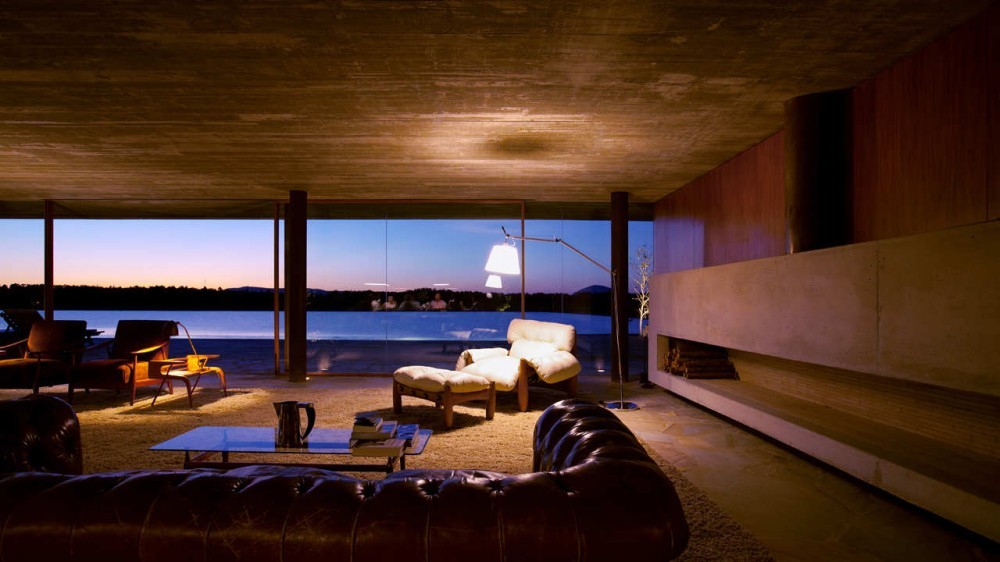 Lounge with leather sofa, armchairs and a long concrete fireplace facing glass walls and a lake at dusk