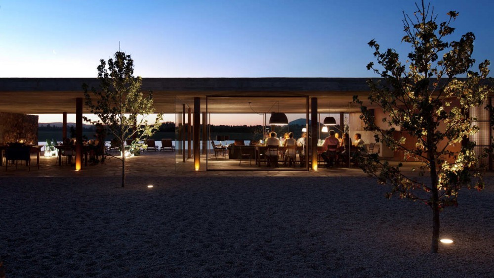 Glazed pavilion with diners inside, seen across a gravel court with two small lit trees at twilight