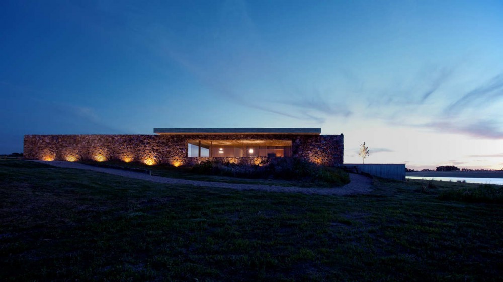 Low stone-walled pavilion with a thin flat roof, uplit along its base, on a grassy rise above the water at dusk