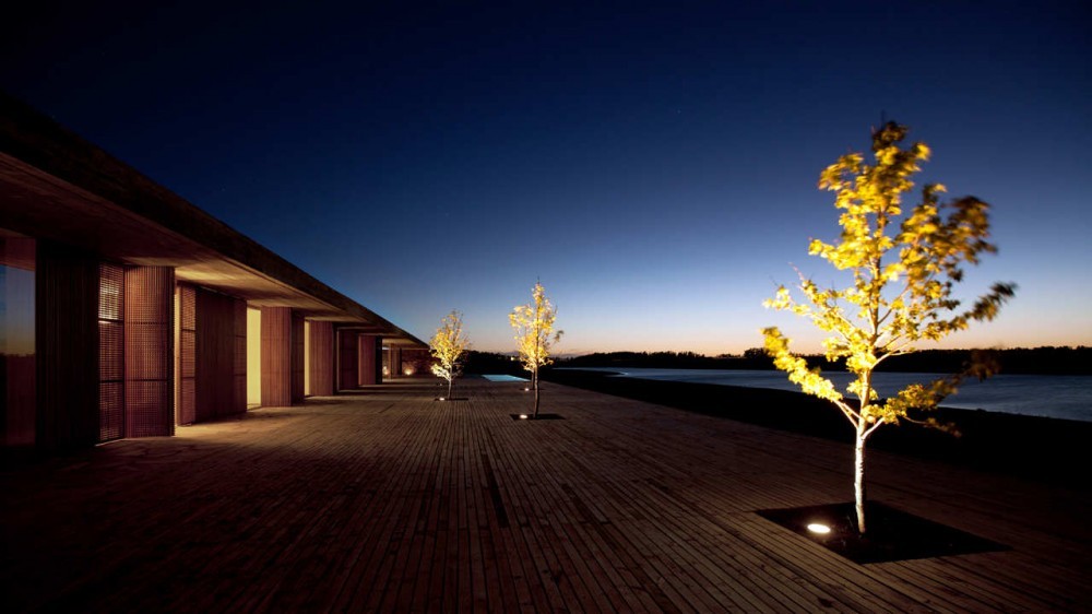 Timber deck running along a shuttered facade, with uplit trees in square inset planters beside the lake under a deep blue sky