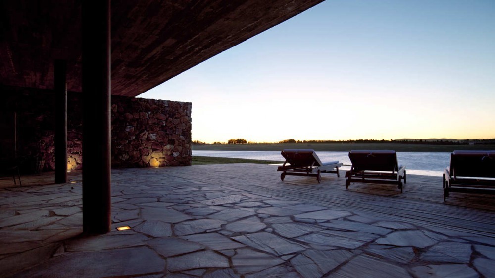 Covered flagstone terrace with steel column, stone wall and three lounge chairs facing the lake at sunset