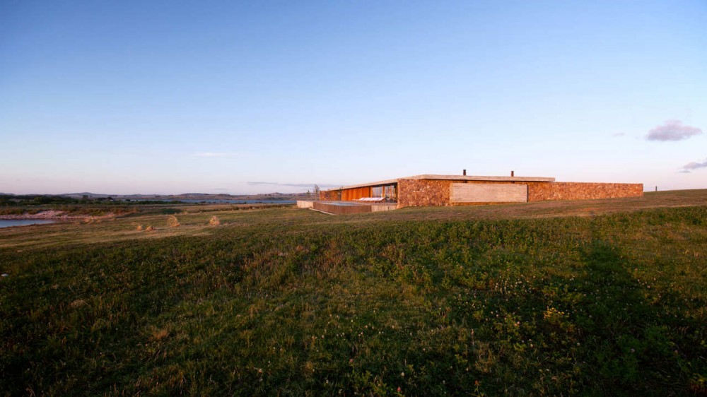 Low stone-walled house with a flat concrete roof set on a grassy rise above a lake at sunset