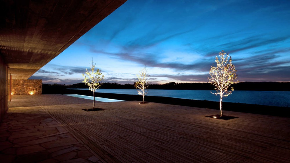 Timber deck with three lit young trees and a pool edge facing the lake under a dusk sky