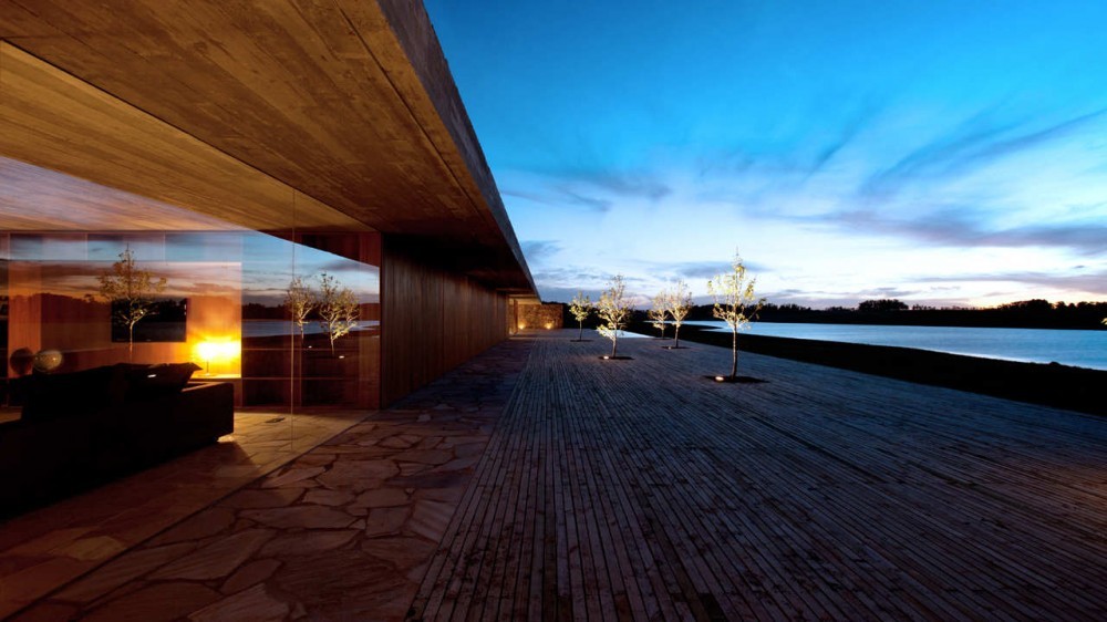 Long cantilevered concrete roof over a glazed living room and flagstone terrace, with lit trees along the deck at dusk