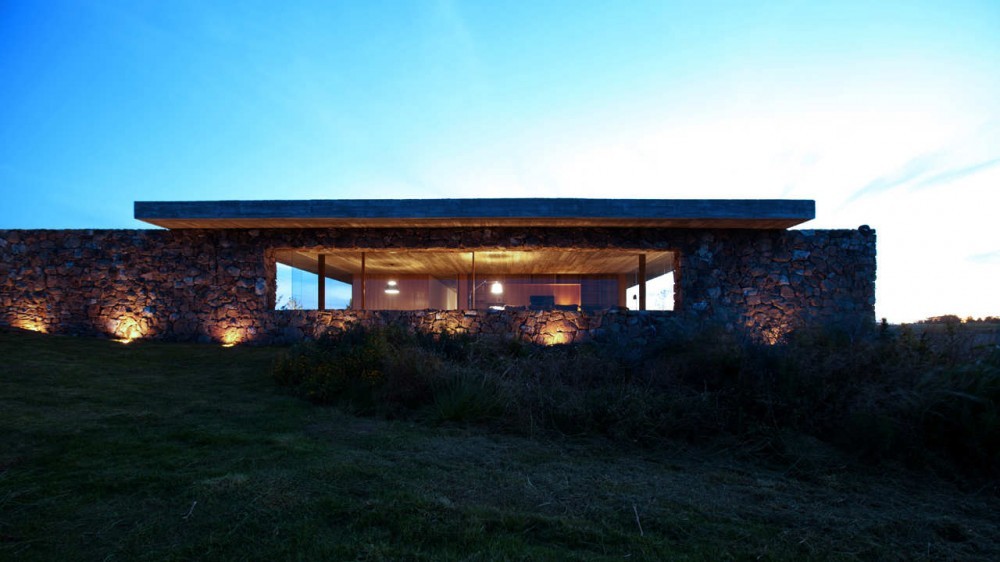Rough stone facade with a wide glazed opening under a thin concrete roof slab, uplit at twilight