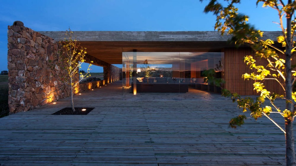 Timber deck with a recessed tree planter running along a rubble stone wall and glazed pavilion, lit at dusk