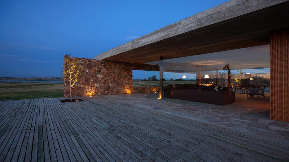 Glass-walled living room under a board-formed concrete roof, with a stone wall and lit sapling on the deck at twilight