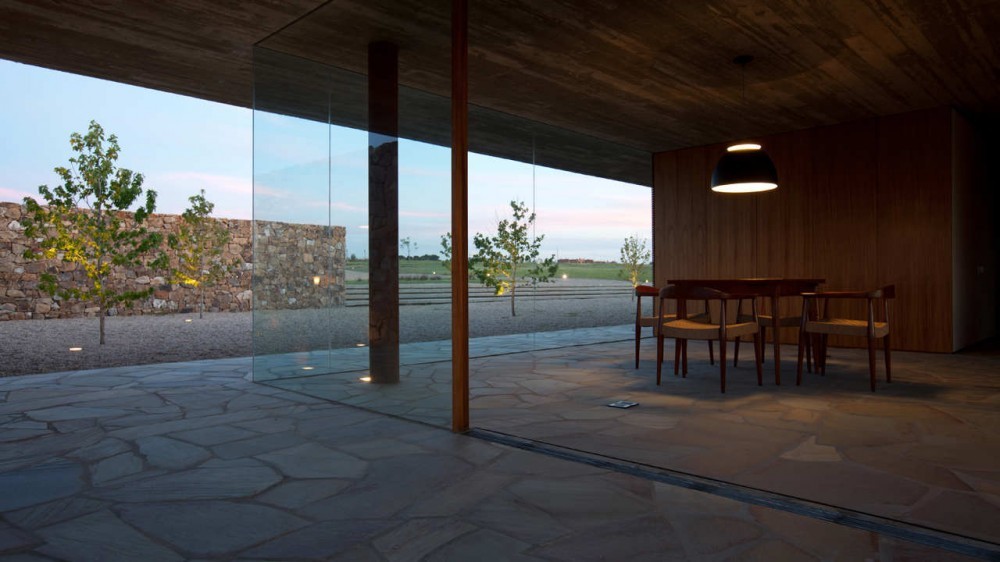 Interior dining area with wood-panelled wall and pendant lamp, looking through floor-to-ceiling glass to a stone wall and young trees