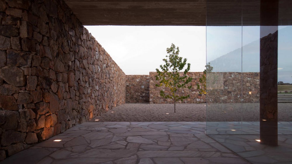 Gravel courtyard framed by rubble stone walls and a single tree, seen through glass from a flagstone floor at dusk
