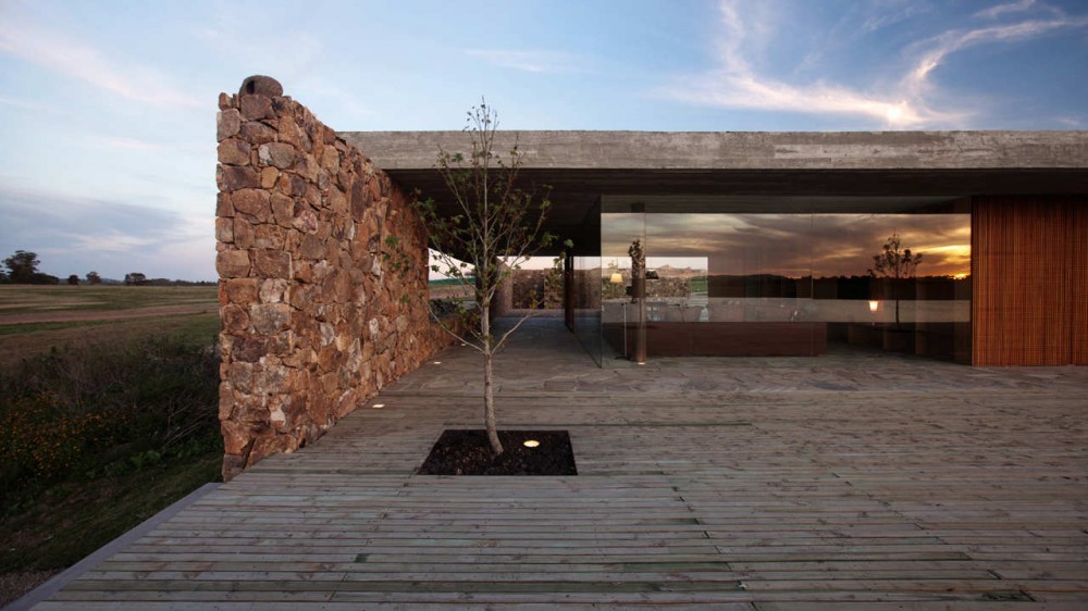 Rubble stone wall meeting a concrete roof slab and glass wall, with a sapling in a deck planter under a cloudy sunset