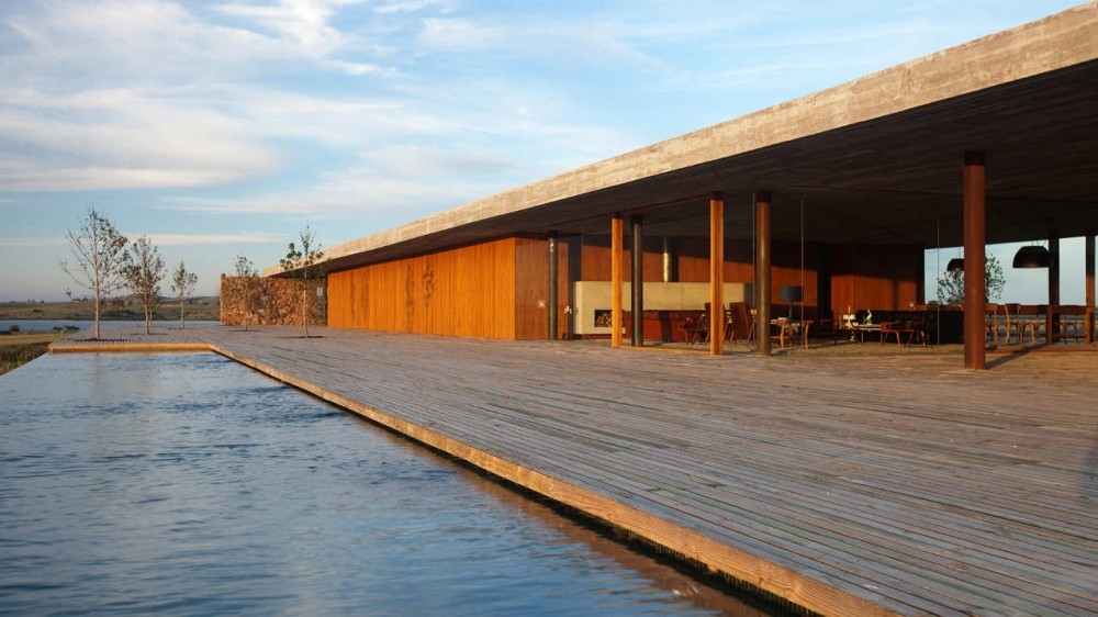 Timber deck beside a long reflecting pool, with a flat concrete roof over a covered pavilion in low evening sun