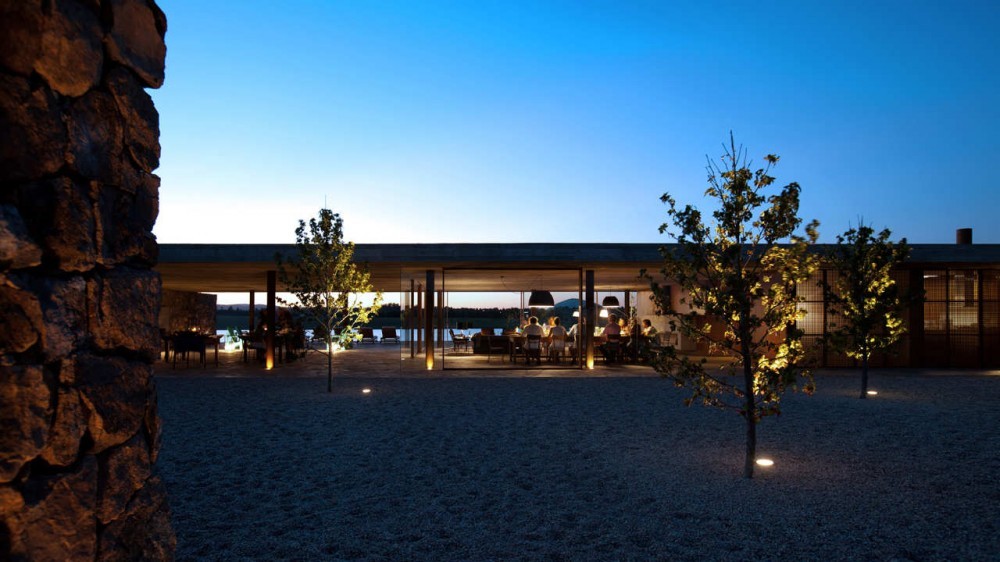 Dusk view across a gravel courtyard with uplit young trees, a rough stone wall and diners in the glazed pavilion