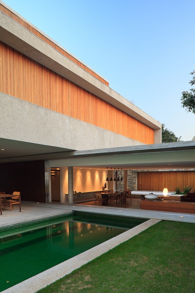 Cantilevered concrete volume with vertical timber slats above a glass-walled dining pavilion and green-tiled pool in clear dusk light