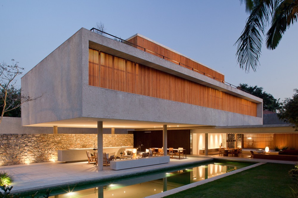 Concrete box on slim columns above a stone wall, lit terrace and long pool at twilight with a palm frond overhead