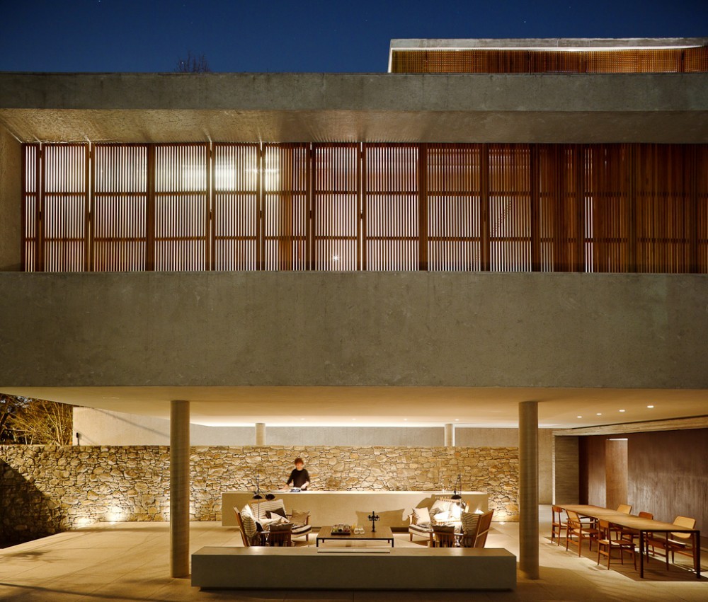 Frontal view of the slatted timber screen over a concrete band, with an outdoor lounge and long dining table below at night