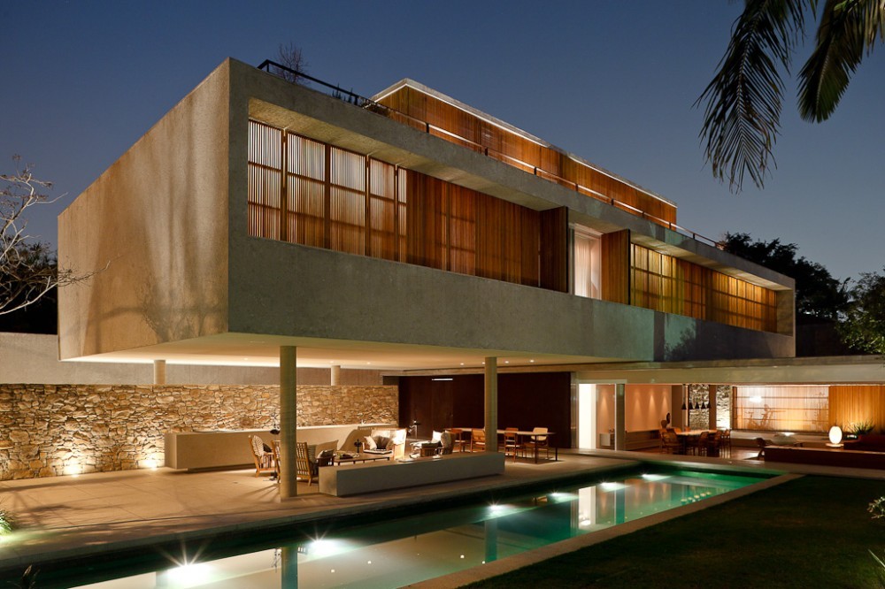 Night view of the raised concrete volume with glowing timber shutters, stone wall and underwater-lit pool reflecting the terrace