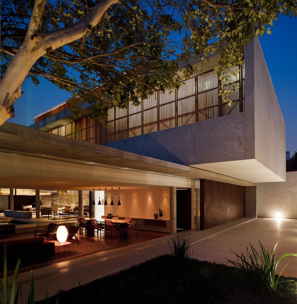 Corner of the concrete upper volume seen through a large tree branch, with a lit living room and dining table below at dusk