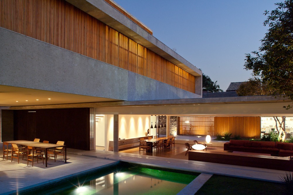 Open ground-floor living area with dining table, red sofa and timber panels beside the pool, under the overhanging concrete volume at dusk