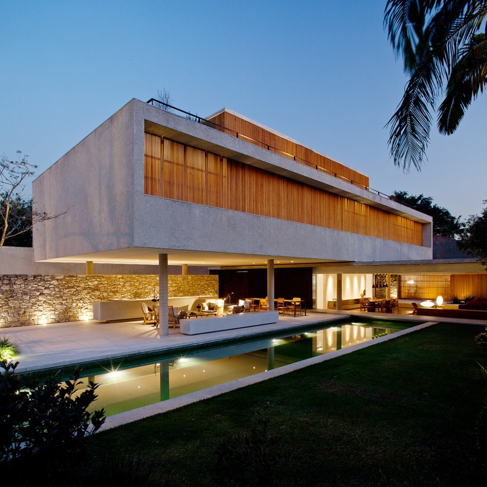 Cantilevered concrete box with timber slat screen above a lit stone wall and lap pool at dusk, palm frond in foreground