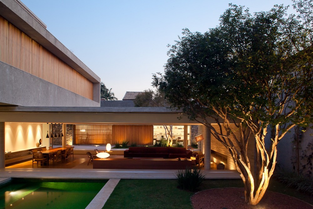 Open living pavilion beneath a concrete slab, with a lit multi-trunk tree in the garden courtyard at twilight