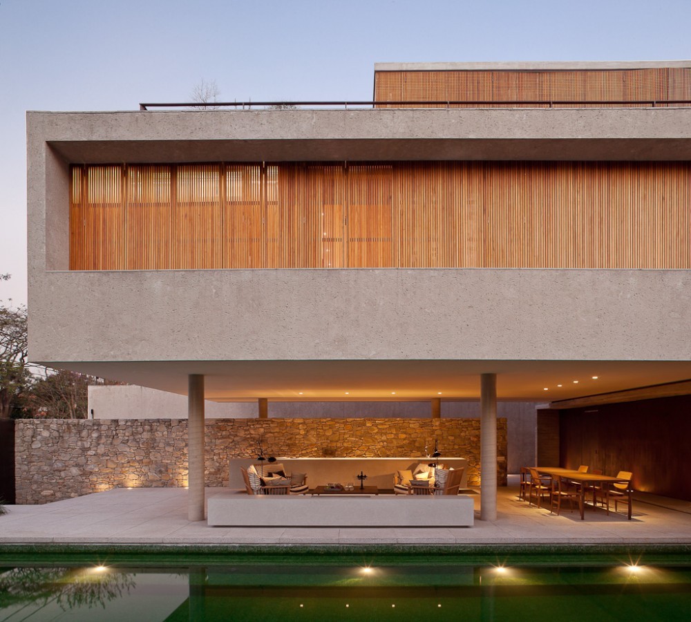 Front elevation of the raised concrete volume with timber slats, two round columns and a lit stone wall above the pool at dusk