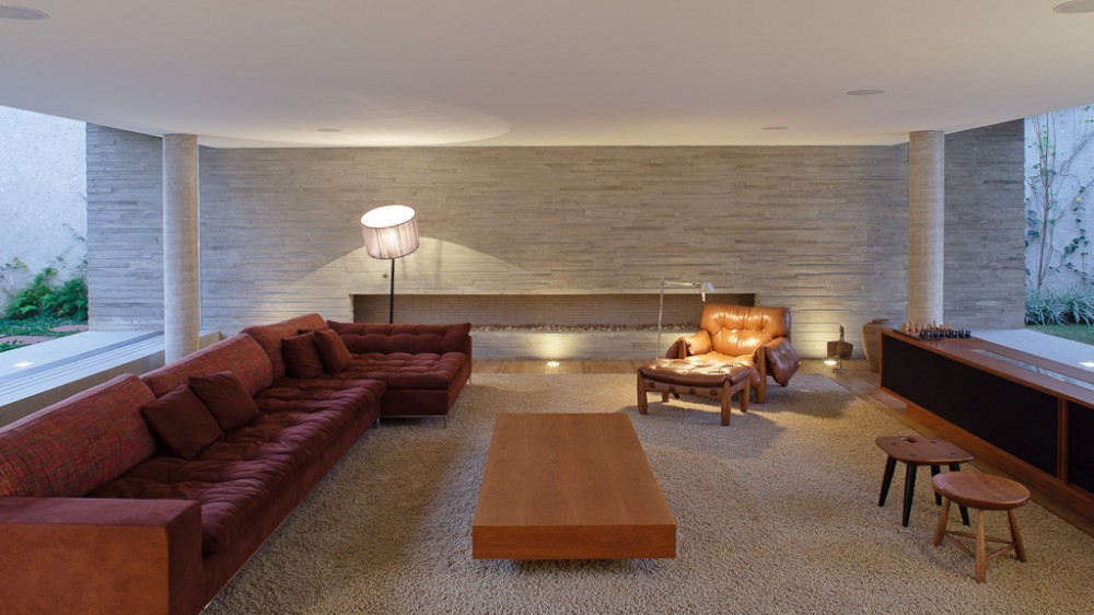 Living room with a rust-red sectional, leather armchair and low wooden table on a shag rug before a board-formed concrete wall