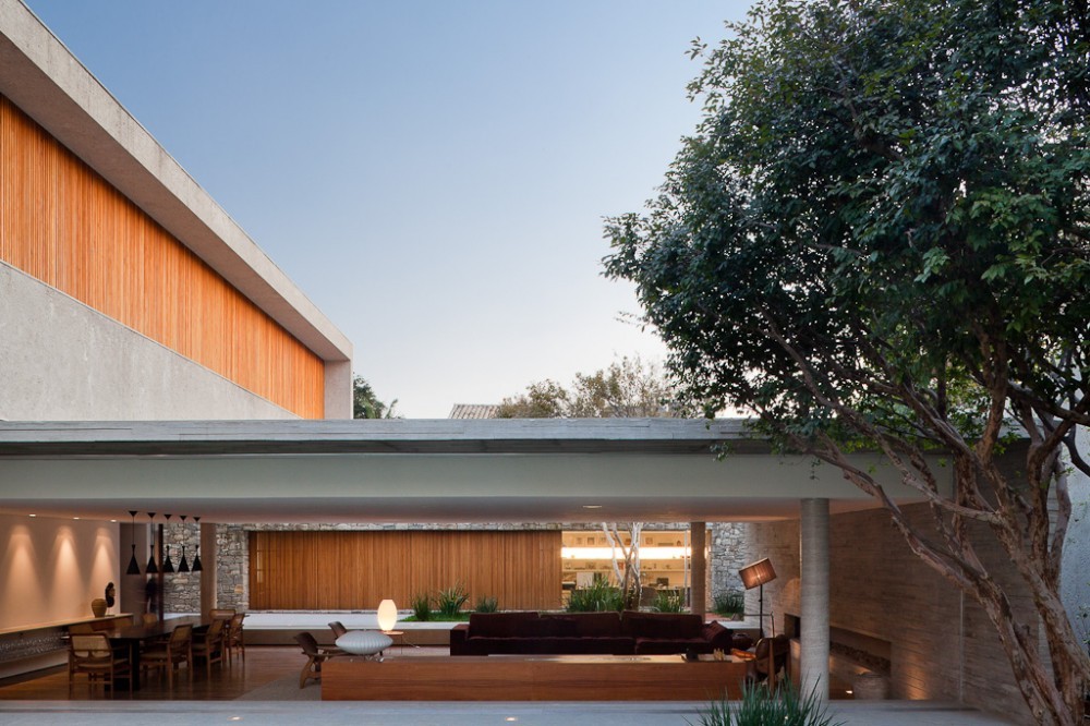 Courtyard view across the open living pavilion toward a timber panel wall, with a leafy tree at right in evening light