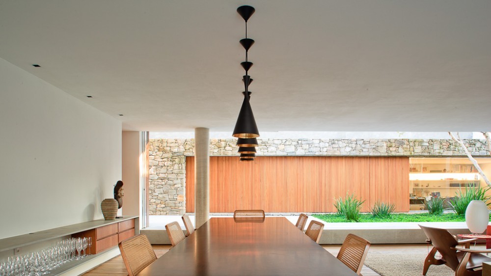 Long wooden dining table under a cluster of black pendant lamps, facing a stone and timber wall and a planted bed