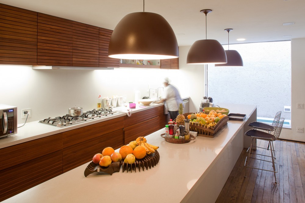 Kitchen with slatted timber cabinets, a long white island holding fruit baskets and three brown dome pendants, with a cook at the counter