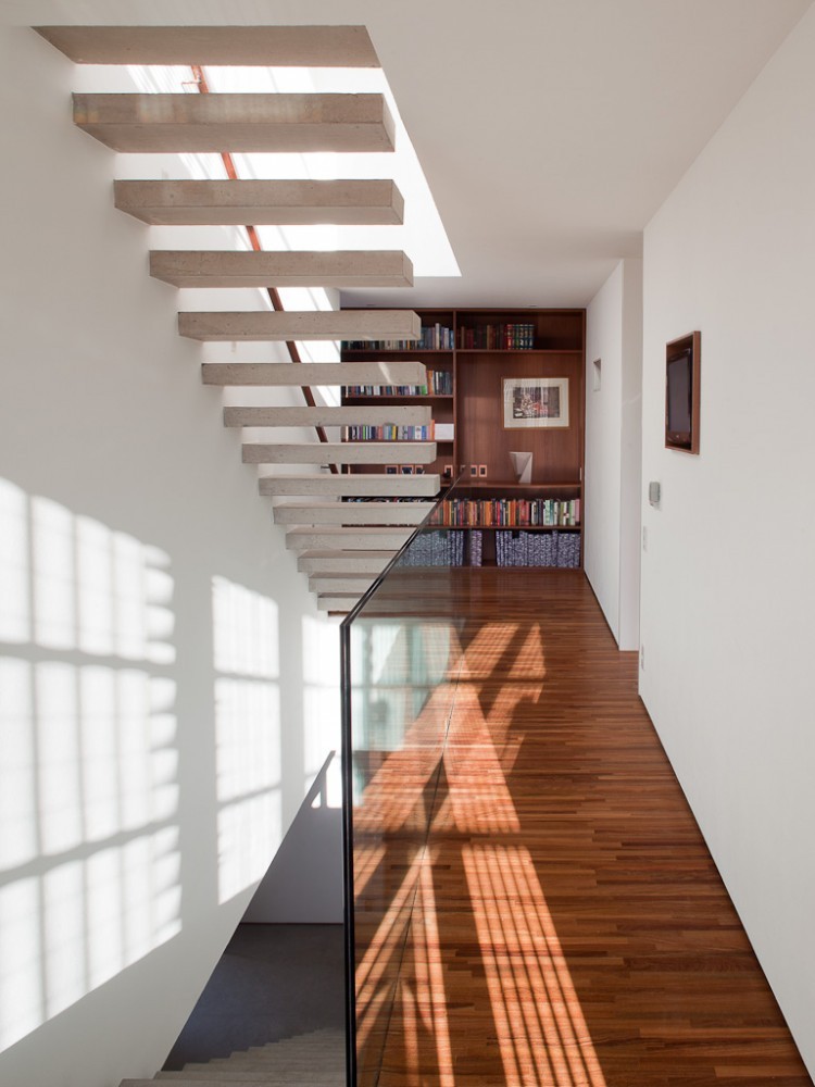 Upper hallway with floating concrete stair treads, a glass balustrade and a timber bookcase, with window light patterning the walls and floor