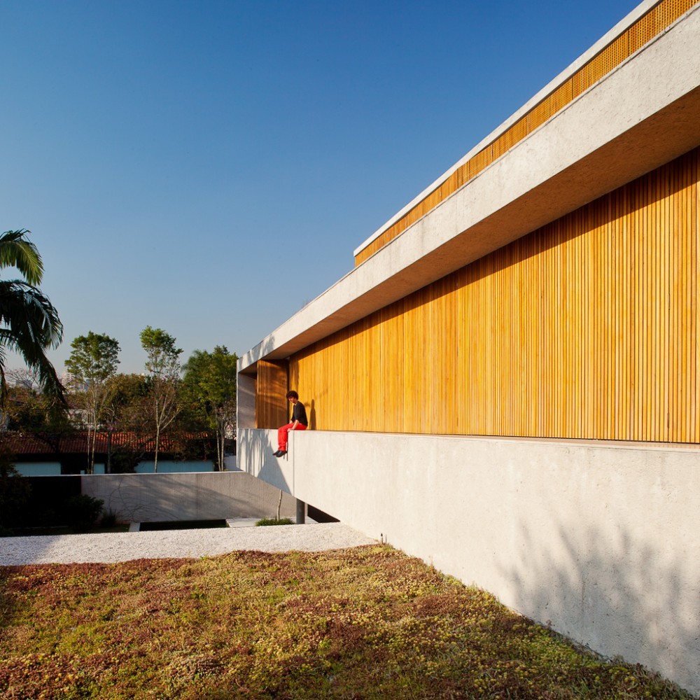 Long concrete and vertical timber slat facade with a person in red trousers sitting on the ledge above a sedum roof