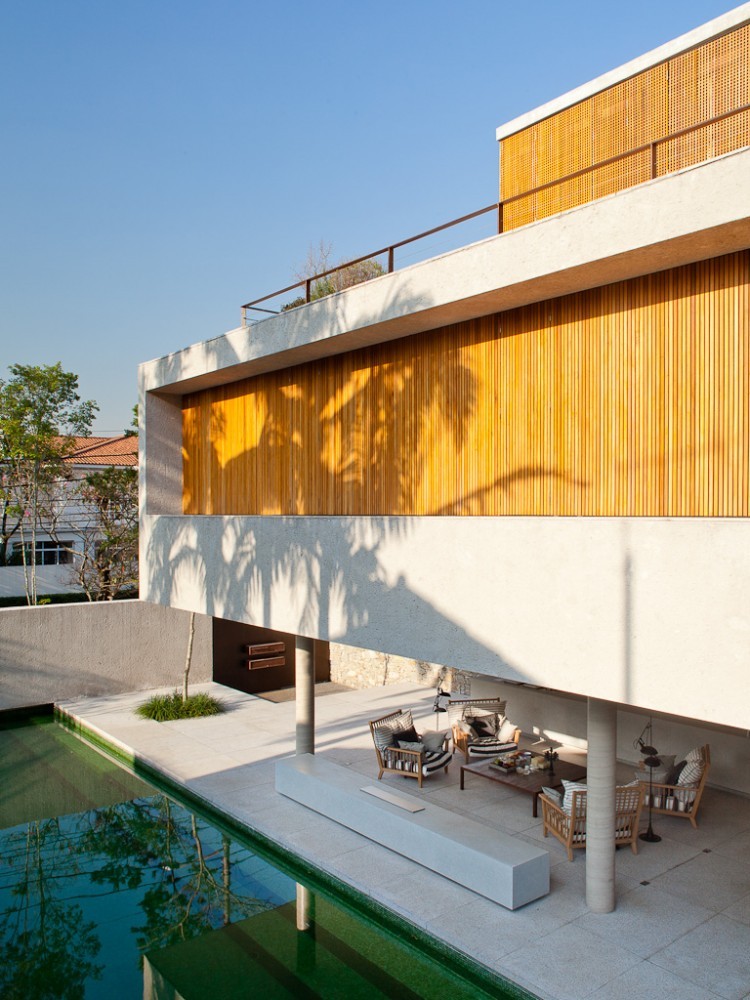 Stacked concrete and timber slat volumes above a covered terrace with armchairs, round columns and a green-tiled pool, with palm shadows on the wall