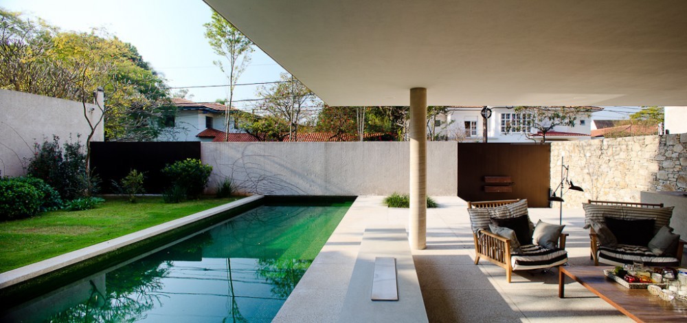 Covered terrace with a round column, wicker armchairs and a long green pool, set against garden walls and neighbouring rooftops in soft daylight