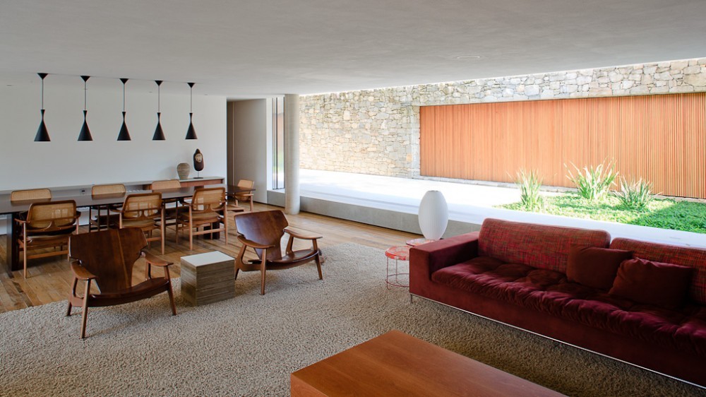 Open living and dining area with pendant lamps, cane-backed chairs and a stone wall with timber slats beyond the glass