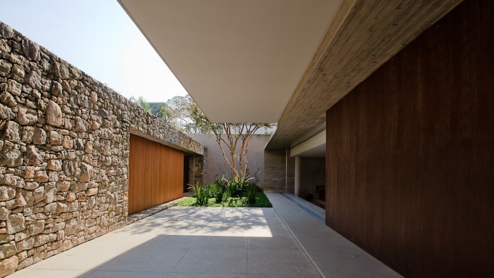 Paved passage between a rough stone wall and a dark timber wall, with a small planted court and tree under a cantilevered slab