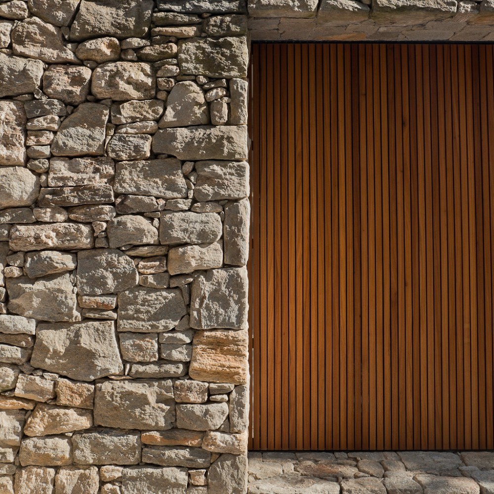 Rough dry-laid stone wall beside a tall vertical timber slat panel in raking sunlight