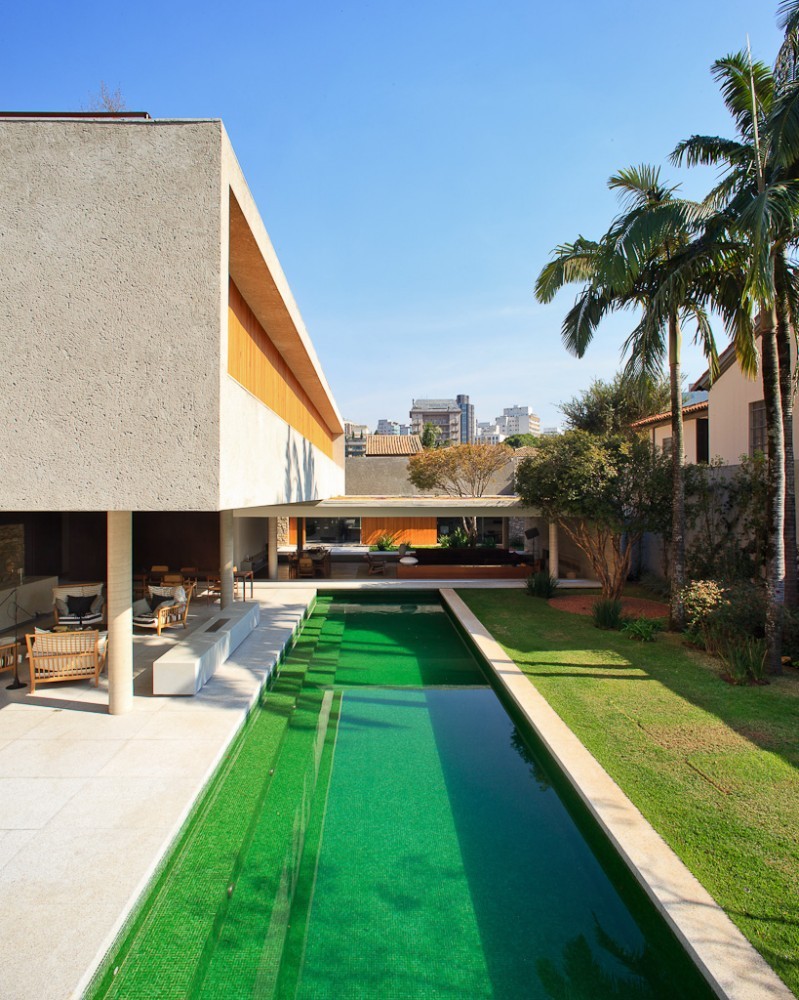 Long green-tiled pool between a raised concrete volume and a lawn, with palm trees under clear blue sky