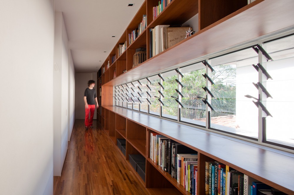 Corridor lined with built-in timber bookshelves and louvered windows, a person in red trousers walking toward the far end