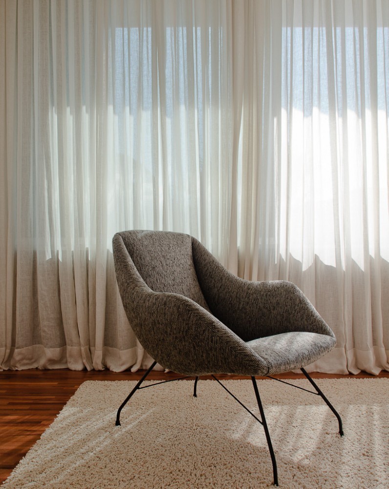 Grey upholstered armchair on thin black legs and a shaggy rug in front of backlit linen curtains