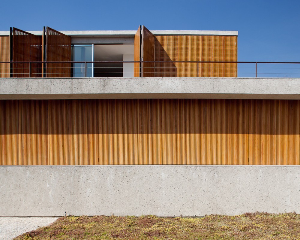 Facade of stacked concrete bands and vertical timber slats, with perforated wooden shutters folded open on the upper level under clear blue sky