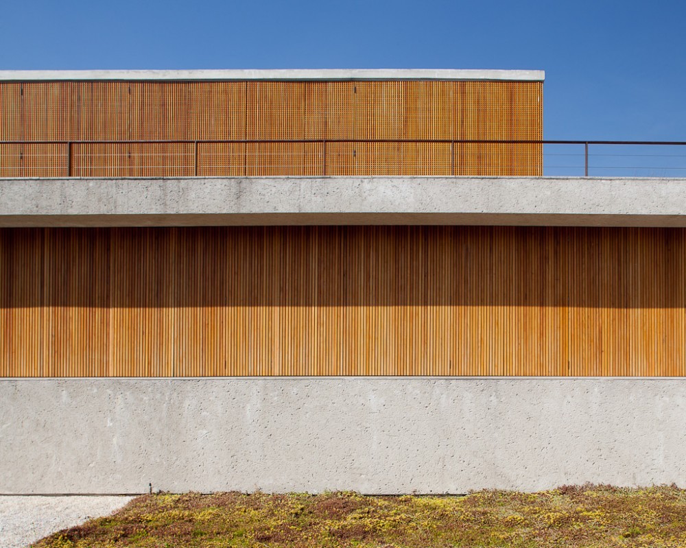 Facade of concrete bands and vertical timber slats with the upper perforated shutters closed, above a low planted roof in bright sun