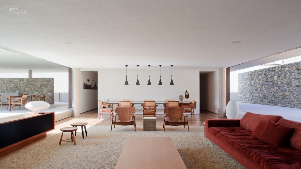 Open living room with a red tufted sofa, wooden armchairs and a long dining table, framed by stone walls in daylight