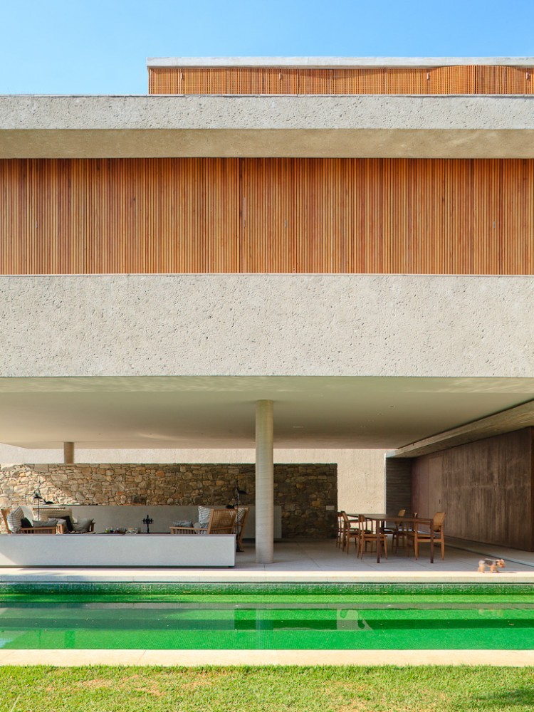 Rear elevation with a concrete volume raised on a single column above a covered terrace, green-tiled pool and a small dog on the lawn